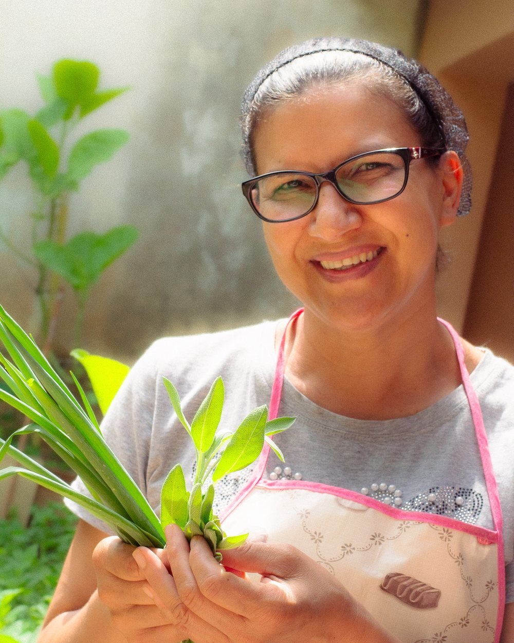 Ná, da Ná Que Faz, sorrindo no quintal com ervas frescas colhidas na mão